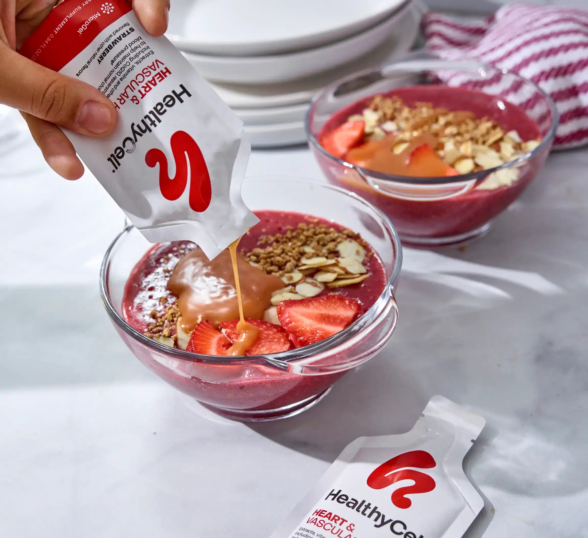 Hand pouring a Healthy Care product into a bowl of fruit and granola, with another bowl and packet in the background.