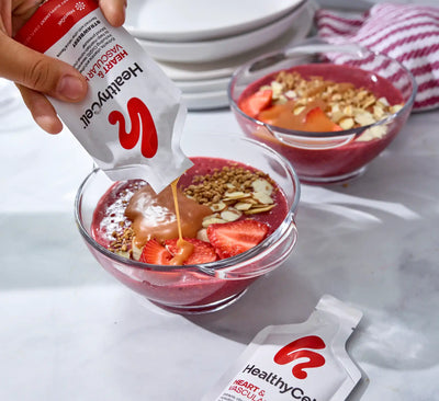 Hand pouring a Healthy Care product into a bowl of fruit and granola, with another bowl and packet in the background.