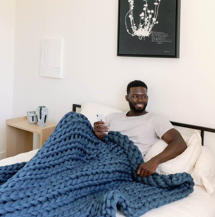 Nuzzie knit weighted blanket in Dusty Blue, draped over a person relaxing in bed