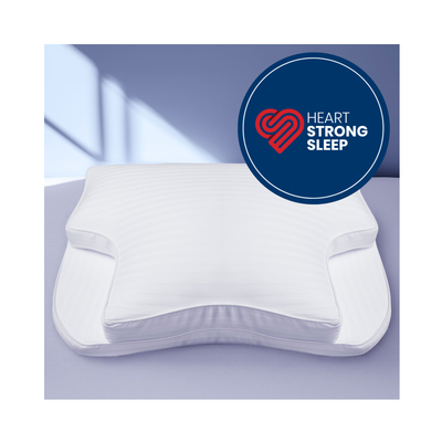 A white contoured support pillow shown on a bed against a soft lavender-blue background, with a circular badge reading 'Heart Strong Sleep' in the upper right.