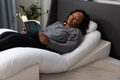 Woman resting comfortably elevated in bed while reading with the Sleep Again Pillow System